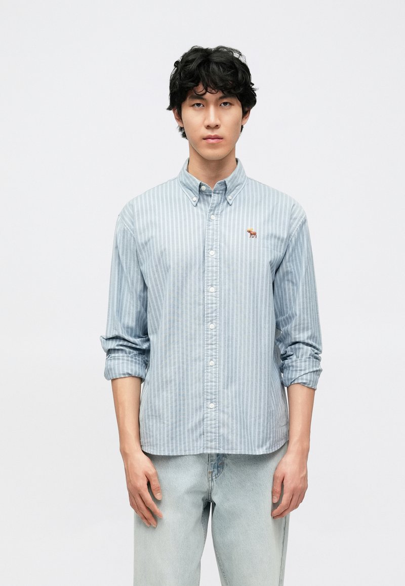 Φωτεινό γαλάζιο πουκάμισο με ρίγες και κουμπιά, κατασκευασμένο από βαμβάκι, με μικρή κεντημένη λεπτομέρεια. Μανίκια γυρισμένα, σε συνδυασμό με ανοιχτόχρωμο τζιν.