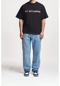 Camiseta negra de algodón con texto blanco "2Y STUDIOS", combinada con vaqueros de mezclilla azul claro de corte relajado y zapatillas negras y blancas.