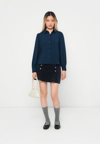 Cămașă bleumarin cu mâneci lungi, asortată cu o fustă mini din corduroy închis, cu butoane argintii, șosete gri până la genunchi și o geantă albă.