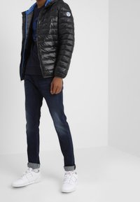 Svart puffjacka med en blå huva, dragkedjestängning och en logotypapplikation. Bärs med mörka jeans och vita sneakers. Slät textur.