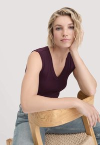 V-Narben, ärmelloses Top aus dunkelviolettem Stoff, kombiniert mit blauen Jeans. Die Person ruht die Hand auf einem hellen Holzstuhl mit geflochtenem Sitz.