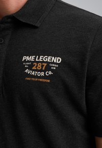 Zwarte poloshirt met een gemêleerde afwerking, met witte en bruine geborduurde tekst: "PME LEGEND," "Flight No. 287," en "Find Your Freedom."