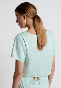 Blusa de manga corta en verde menta con un sutil estampado de leopardo y un dobladillo recto. Cuenta con un detalle de lazo en la parte posterior para un ajuste ajustable.