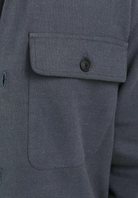 Mörkgrå skjorta med strukturerad tyg, stor bröstficka och en svart knapp. Designen inkluderar en klassisk krage och framknäppning.