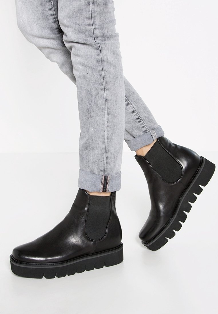 Personne portant un jean gris clair retroussé et des bottines Chelsea en cuir noir avec des semelles épaisses et rainurées sur un sol blanc.