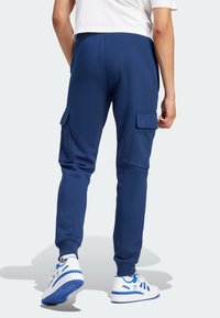 Man i marinblå cargojoggers och vita Adidas-sneakers med blå accenter, som står mot en ljus bakgrund.