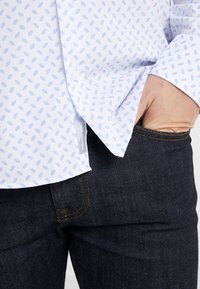 Vit skjorta med små ljusblå mönstrade prickar, långa ärmar och en etikett, kombinerad med mörka denimjeans. Tyget verkar slätt.