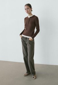 Woman wearing a brown long-sleeve knit top over a white shirt, gray wide-leg jeans, and beige shoes with black spots, standing with hands in pockets.