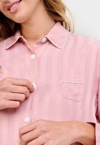 Mujer sonriendo mientras abotona una camisa de pijama a rayas en rosa claro con ribetes blancos en el collar y el bolsillo.
