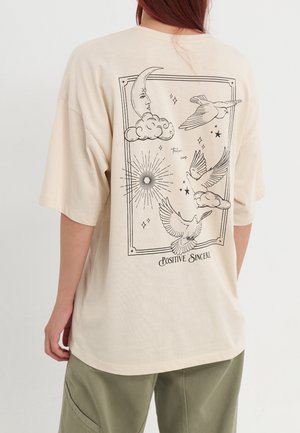 Persona que lleva una camiseta beige con dibujo lineal negro de luna, nubes, estrellas, palomas y las palabras "Positive Sincere" en la parte trasera.