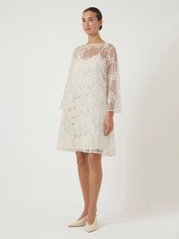 Γυναίκα με λευκό φόρεμα μέχρι το γόνατο, με διαφανή μακριά μανίκια και κεντήματα από λουλούδια με παγιέτες, συνδυασμένο με κρεμ ψηλοτάκουνα παπούτσια.