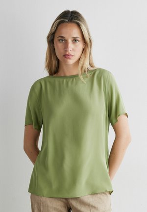 Junge Frau mit blondem Haar trägt eine schlichte hellgrüne Kurzarmbluse und beige Hosen vor einem weißen Wandhintergrund.