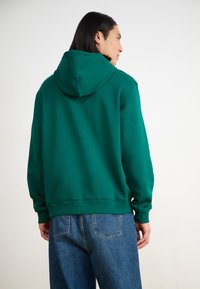 Hanorac verde din bumbac cu glugă ajustabilă, manșete și tiv din tricot. Purtați-l cu blugi din denim albastri, evidențiind un stil casual și relaxat.