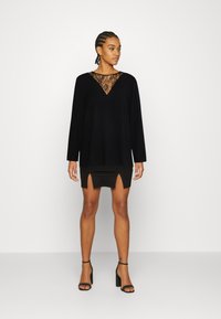 Blusa longa preta com detalhe em renda no decote em V, combinada com um vestido preto justo com fendas laterais e sapatos de salto com tira no tornozelo.