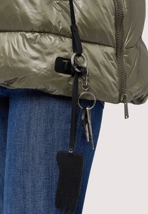 Chiavi attaccate a un portachiavi a forma di guanto nero, appeso a una giacca a vento verde oliva indossata sopra jeans blu.