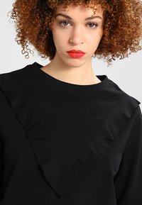 Fekete, hosszú ujjú felső, V-alakú fodor részlettel a nyakvonalnál, puha anyagból, sima textúrával.