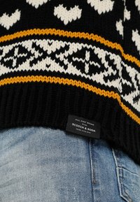 Maglione nero lavorato a maglia con motivi geometrici bianchi e gialli, polsini a costine e un'etichetta con la scritta "SCOTCH & SODA" sull'orlo.