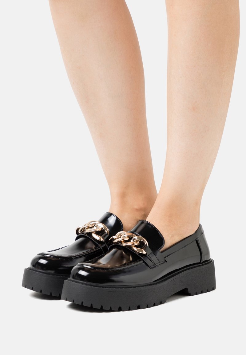 Mocassins en cuir verni noir avec une semelle plateforme épaisse, ornés d'une chaîne dorée sur le dessus. Texture lisse avec une forme arrondie.