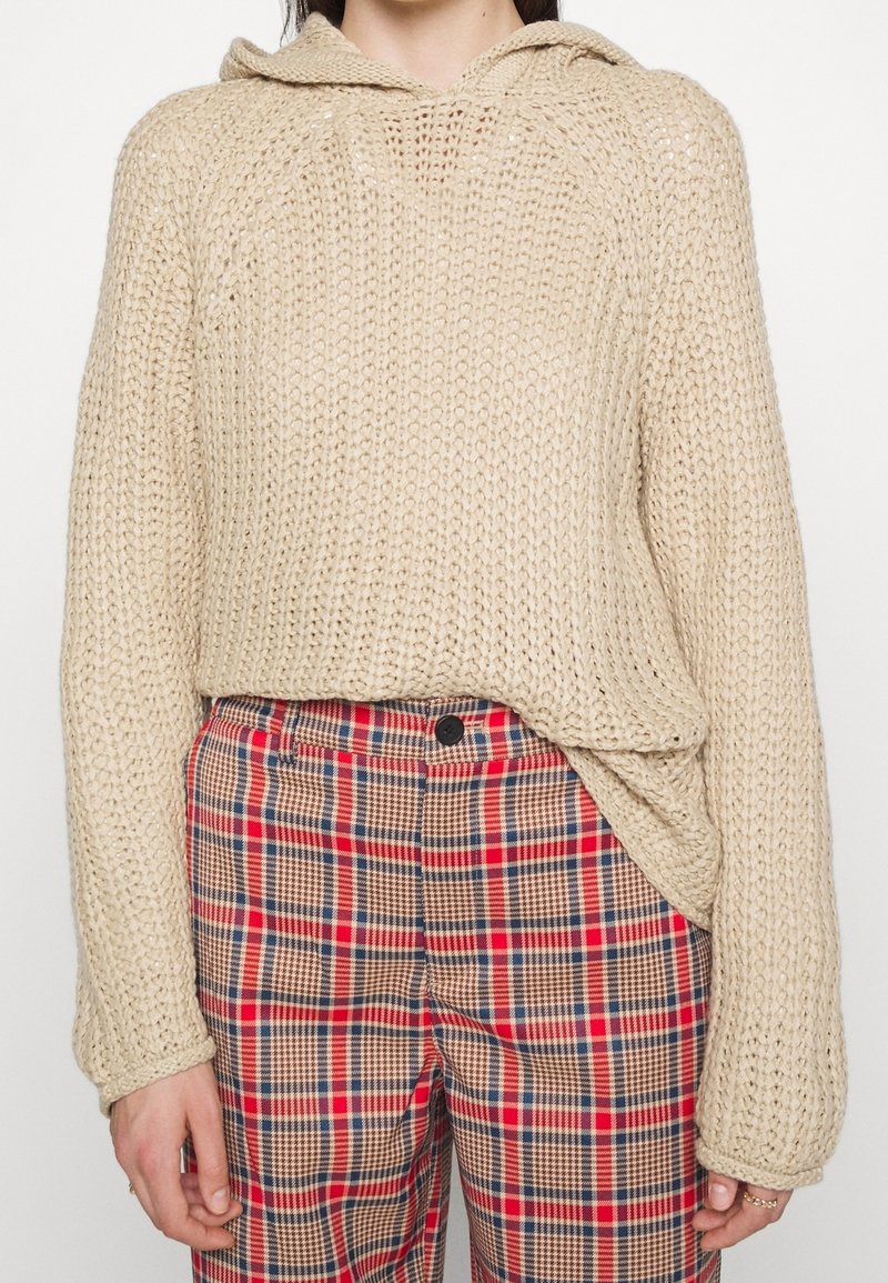 Beige stickad hoodie med lös passform och vida ärmar, tillsammans med rutiga byxor med mönster i röd, blå och brun.