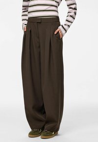 Tumed pruunid laiade säärtega püksid kõrge vöökohaga, volditud disainiga ja küljetaskutega; kantud triibulise pika varrukaga pluusi ja roheliste jalanõudega.