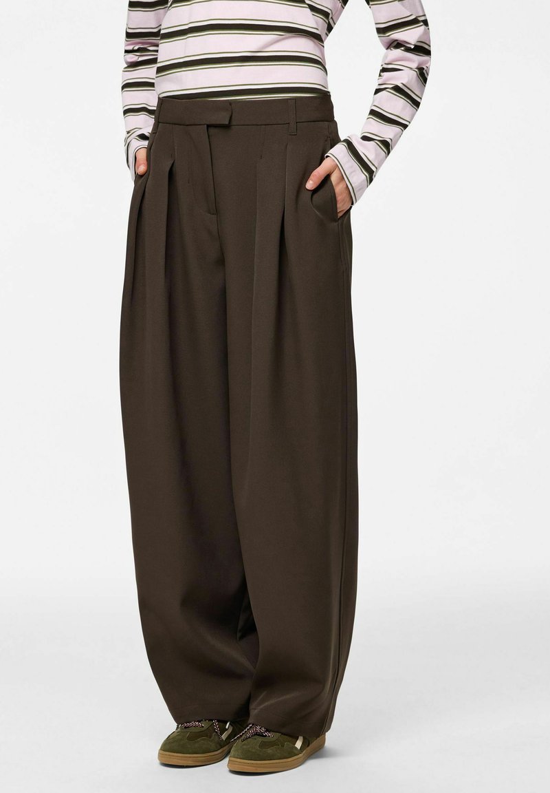 Tumed pruunid laiade säärtega püksid kõrge vöökohaga, volditud disainiga ja küljetaskutega; kantud triibulise pika varrukaga pluusi ja roheliste jalanõudega.