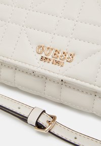 Cremefarvet quiltet håndtaske med et guld "GUESS" logo og "EST 1981" detalje. Den har en smal rem med en guld spænde og en glat tekstur.