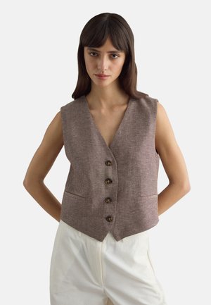 Frau mit langen dunklen Haaren trägt eine braune ärmellose Weste mit Fischgrätmuster und weiße, hochtaillierte Hosen, steht mit den Händen hinter dem Rücken.