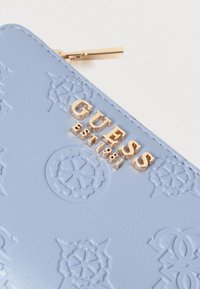 Portofel din piele albastru deschis, cu modele florale embossate și logo-ul "GUESS EST. 1981" în ton auriu. Închidere cu fermoar și accesorii aurii.