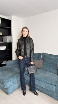 Casaco de couro preto com fecho de correr, usado sobre uma gola alta preta, combinado com jeans flare escuros e uma pequena carteira preta.