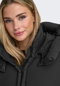 Musta toppatakki, jossa on korkea kaulus ja vetoketjusuljenta. Materiaali näyttää teksturoidulta ja siinä on tikattuja osioita. Malli esittää neutraalia ilmettä.