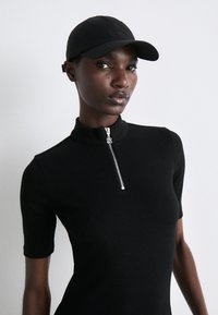 Černé upnuté šaty s krátkými rukávy a vysokým límečkem, obsahující stříbrný zip na krku. Model nosí shodný černý klobouk.
