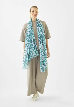 Lachende vrouw staande, gekleed in een oversized beige outfit en witte sneakers, met een lange blauw met patroon sjaal om haar nek gedrapeerd.