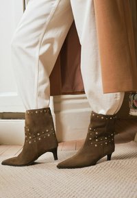 Hnedé semišové členkové topánky s špicatou špičkou a nízkym podpätkom, s dekoračnými zlatými nitmi usporiadanými v geometrickom vzore.