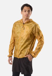 Jeune homme portant une veste à capuche au motif camouflage jaune et un short noir, la main gauche dans la poche, debout devant un fond blanc.