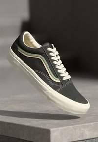 Black low-top sneaker with white sole, white laces, and a green and beige side stripe hovering above a concrete ledge.