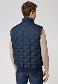 Navyblaue Steppweste mit einem gesteppte Muster, hohem Kragen und elastischem Saum, kombiniert mit einem beigefarbenen Langarmshirt und dunklen Jeans.