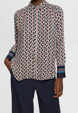 Woman wearing a long-sleeve patterned blouse with geometric shapes in navy, white, and rust, paired with dark navy pants.