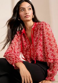 Blusa rossa a fantasia con tessuto traslucido, maniche arricciate, scollo a V con dettagli a laccio, abbinata a pantaloni neri.