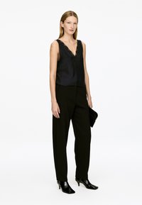 Woman standing wearing a black lace-trimmed sleeveless top, black trousers, black heeled boots, and holding a black clutch against white background.