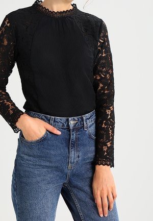 Sort top med plisseret front og lange ærmer i sort blonde med bølgede kanter, parret med højtaljede blå denim jeans.