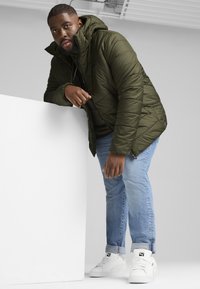 Giubbotto imbottito verde oliva con cappuccio, chiusura a zip e design trapuntato, abbinato a jeans azzurri chiari e scarpe da ginnastica bianche.