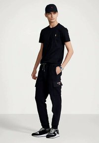 Jauns vīrietis, tērpies melnā cepurē, melnā T-kreklā, melnās kraukšķīgās biksēs un melnbaltās kedās, stāv ar vienu roku kabatā.