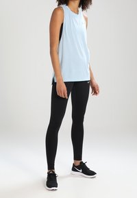 Top atletico senza maniche di colore azzurro chiaro con spacchi laterali, indossato sopra una canotta nera. Combinato con leggings neri e sneakers nere con dettagli bianchi.
