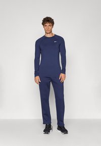 Tricou sportiv cu mâneci lungi, de culoare bleumarin, cu mâneci raglan, asociat cu pantaloni asortați. Ambele sunt fabricate dintr-un material neted și elastic. Pantofi sport negri.