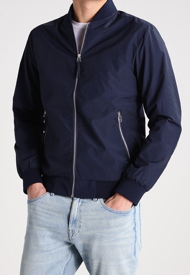 Homme portant une veste bomber bleu marine zippée avec des poches latérales zippées et un jean bleu clair, les mains dans les poches.