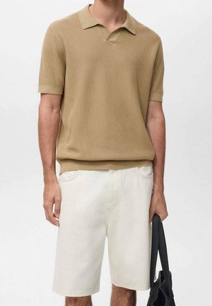 Homme portant un polo en maille beige à manches courtes et un short blanc longueur genou, tenant un sac cabas noir à ses côtés.