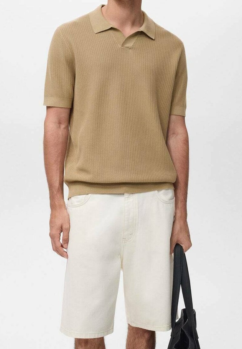 Homme portant un polo en maille beige à manches courtes et un short blanc longueur genou, tenant un sac cabas noir à ses côtés.