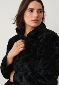 Sort faux fur jakke med en blød tekstur, knaplukning og høj krave. Stoffet har et fyldigt, pelslignende udseende.