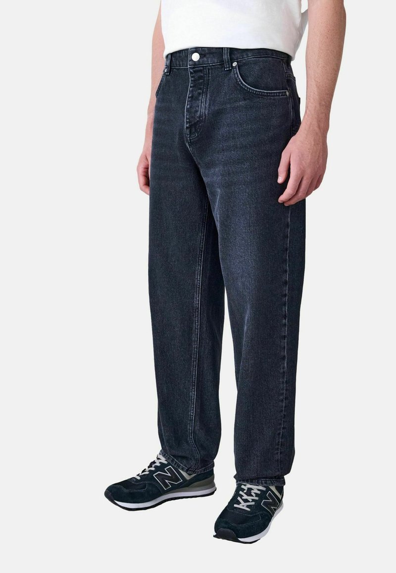 Hombre vestido con jeans de corte relajado de color gris oscuro y zapatillas negras de New Balance, de pie contra un fondo liso.