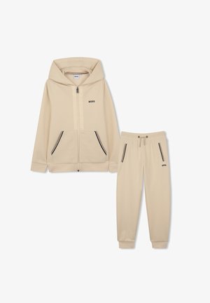 Beige BOSS ritsvest met voorzakken en bijpassende joggingbroek met trekkoord, beide voorzien van zwart-witte ritssluitingaccenten.